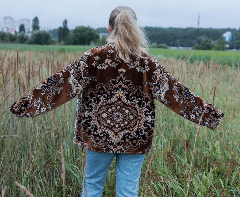 May include: A brown jacket with an ornate, repeating pattern in beige and black. The jacket is worn over blue jeans. The person is standing in a field of tall grass with a background of trees and a cloudy sky.