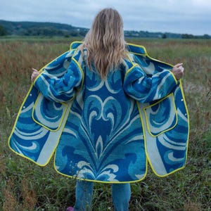 May include: A blue and white patterned coat with yellow edging. The coat has a large floral design and two pockets. The person is standing in a field. The coat is open, showing the interior lining.