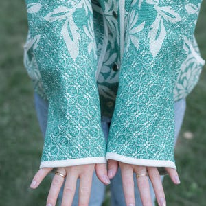 May include: A teal and white patterned jacket featuring a large floral design on the upper portion and a geometric grid pattern on the sleeves. The jacket has a white trim along the front opening and cuffs, with visible silver-toned snap buttons.