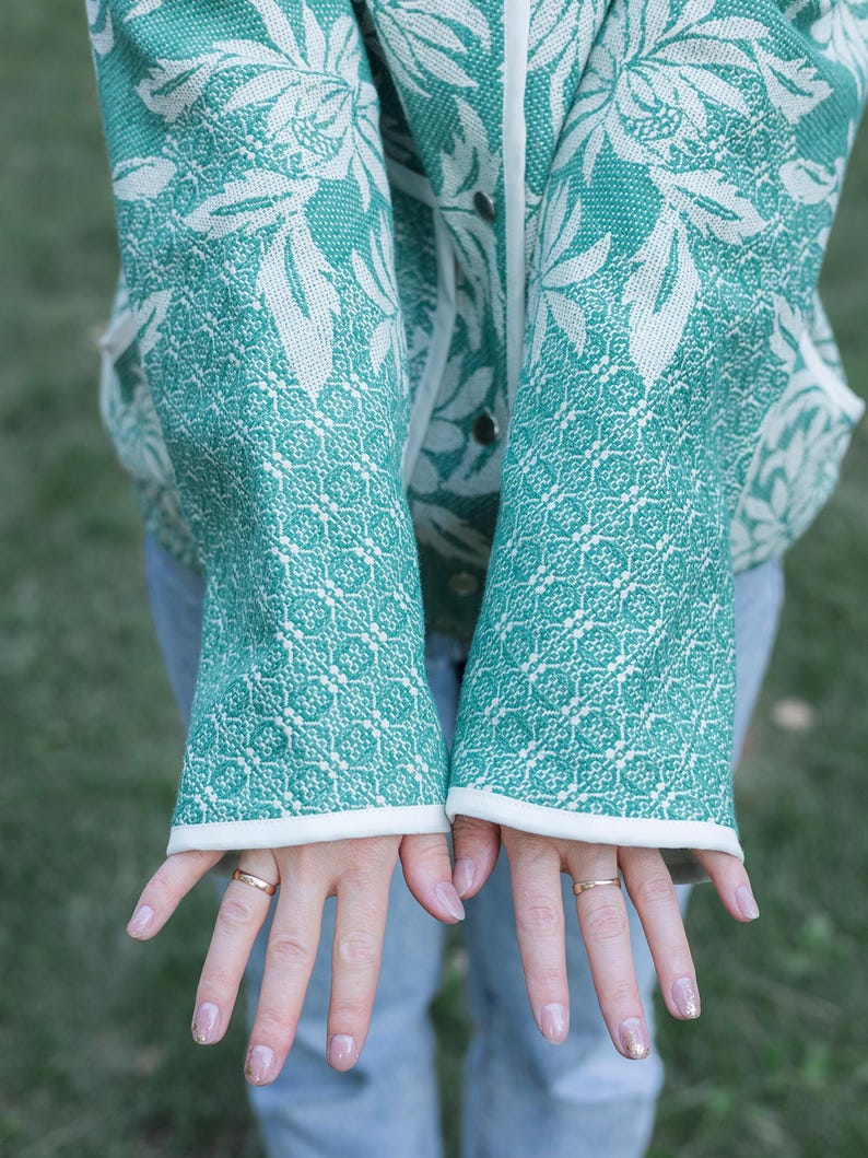 May include: A teal and white patterned jacket featuring a large floral design on the upper portion and a geometric grid pattern on the sleeves. The jacket has a white trim along the front opening and cuffs, with visible silver-toned snap buttons.