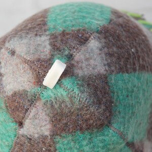 Puede incluir: Primer plano de un sombrero hecho a mano con un patr&oacute;n de cuadros en tonos marr&oacute;n, verde azulado y gris. El sombrero tiene una etiqueta rectangular blanca adjunta. Los bordes est&aacute;n cosidos con hilo verde brillante.