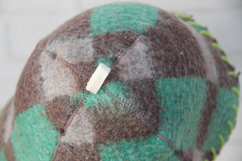 Puede incluir: Primer plano de un sombrero hecho a mano con un patr&oacute;n de cuadros en tonos marr&oacute;n, verde azulado y gris. El sombrero tiene una etiqueta rectangular blanca adjunta. Los bordes est&aacute;n cosidos con hilo verde brillante.