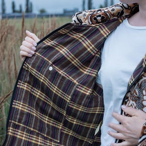 May include: A brown and black patterned jacket with a plaid interior. The jacket has a zipper closure and a decorative collar. The interior features a brown, yellow, and purple plaid pattern. The jacket is being held open, revealing the interior.