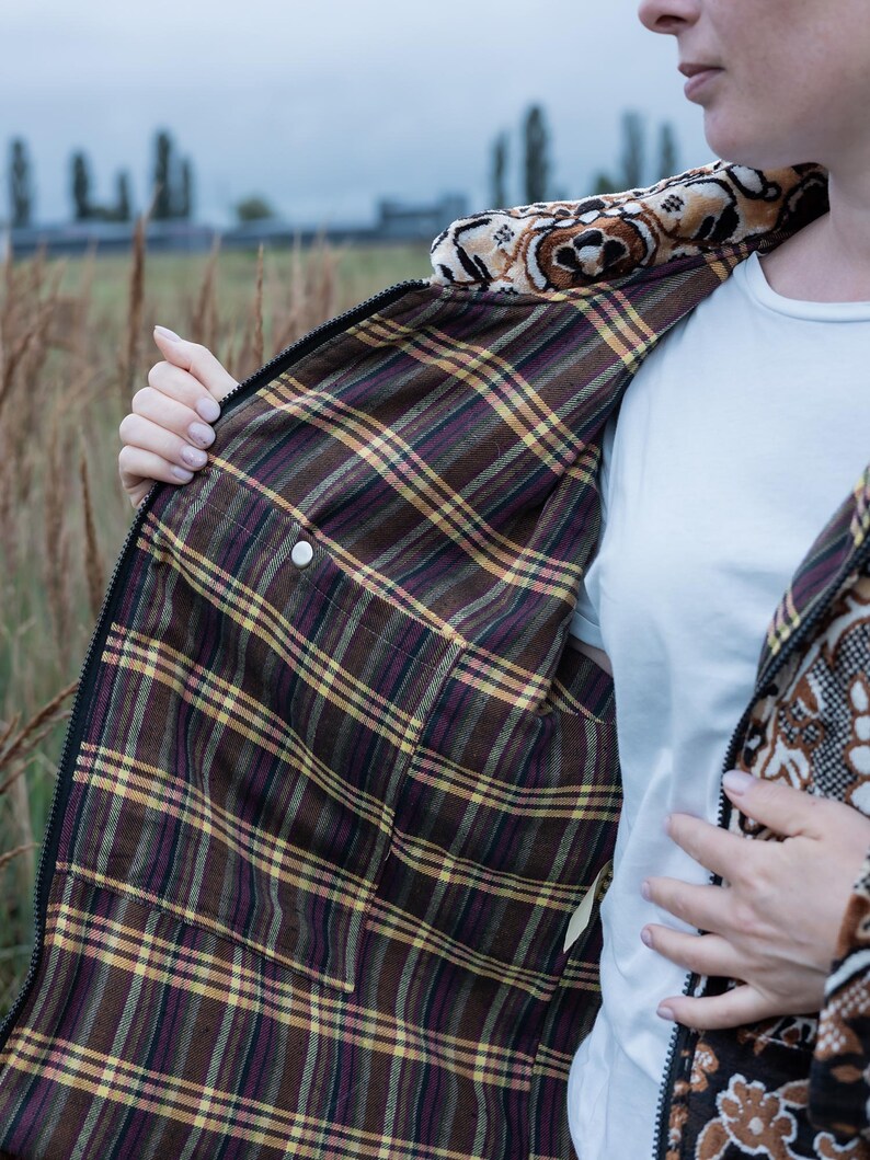May include: A brown and black patterned jacket with a plaid interior. The jacket has a zipper closure and a decorative collar. The interior features a brown, yellow, and purple plaid pattern. The jacket is being held open, revealing the interior.