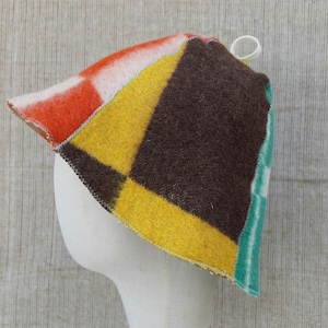 May include: A patchwork hat with a geometric design. The hat features sections of orange, white, yellow, brown, and teal. It is displayed on a white mannequin head. A small white loop is attached at the top. The hat's design is reminiscent of a Mondrian painting.