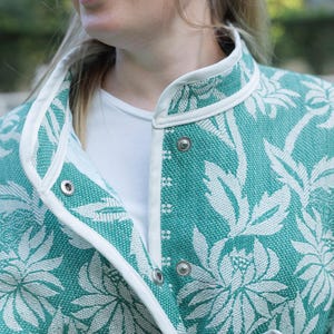 May include: A teal and white floral patterned jacket featuring a high collar, white piping trim along the edges, and silver-toned metal snap button closures. The fabric has a textured weave, and the garment is worn over a plain white shirt.