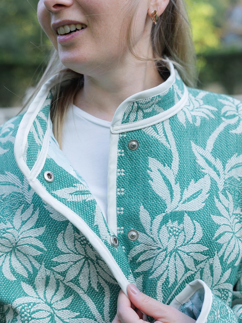 May include: A teal and white floral patterned jacket featuring a high collar, white piping trim along the edges, and silver-toned metal snap button closures. The fabric has a textured weave, and the garment is worn over a plain white shirt.