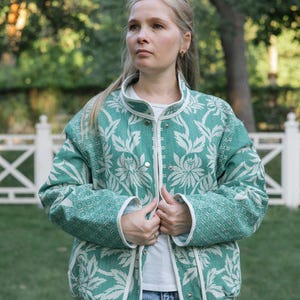 May include: A long-sleeved jacket featuring a teal and white floral pattern with a high collar and silver snap-button closures. The garment is trimmed with white piping along the front opening, cuffs, and hem, and is worn over a white shirt.