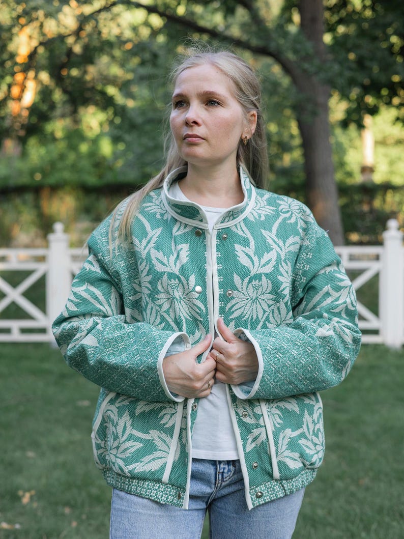 May include: A long-sleeved jacket featuring a teal and white floral pattern with a high collar and silver snap-button closures. The garment is trimmed with white piping along the front opening, cuffs, and hem, and is worn over a white shirt.