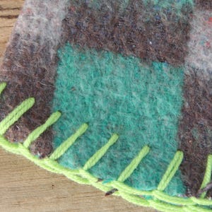 Puede incluir: Primer plano de una secci&oacute;n de una manta con un dise&ntilde;o de patchwork. La manta presenta cuadrados de lana verde azulado, marr&oacute;n y gris. El borde est&aacute; acabado con hilo verde brillante en una puntada decorativa.