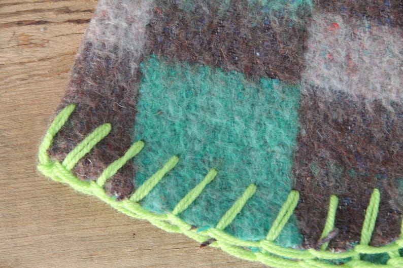 Puede incluir: Primer plano de una secci&oacute;n de una manta con un dise&ntilde;o de patchwork. La manta presenta cuadrados de lana verde azulado, marr&oacute;n y gris. El borde est&aacute; acabado con hilo verde brillante en una puntada decorativa.