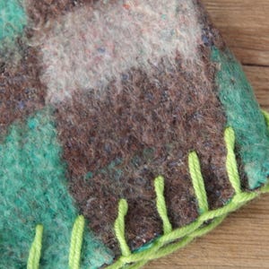Puede incluir: Primer plano de un textil hecho a mano con un patr&oacute;n de cuadros en tonos marr&oacute;n, verde azulado y blanco. El borde est&aacute; acabado con costuras verde brillante, creando una apariencia texturizada y r&uacute;stica. La tela parece ser lana afieltrada.