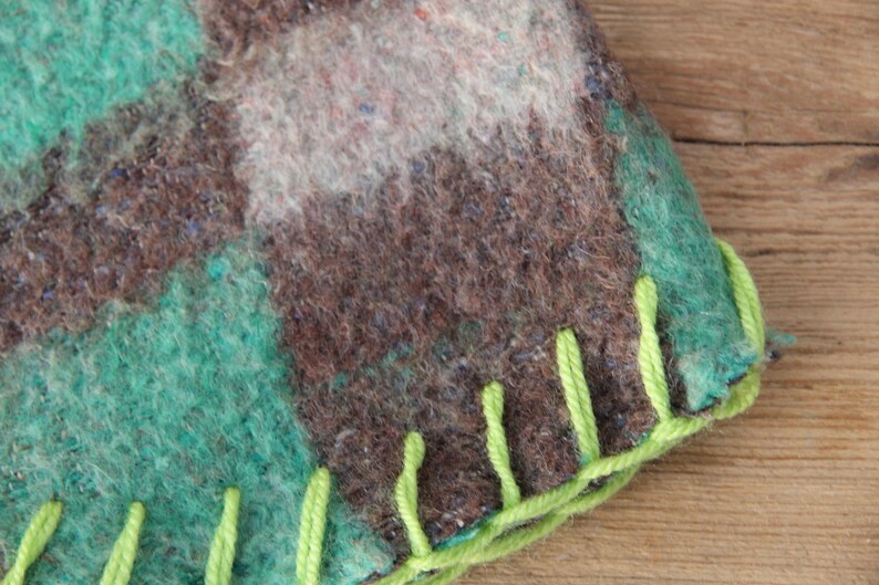 Puede incluir: Primer plano de un textil hecho a mano con un patr&oacute;n de cuadros en tonos marr&oacute;n, verde azulado y blanco. El borde est&aacute; acabado con costuras verde brillante, creando una apariencia texturizada y r&uacute;stica. La tela parece ser lana afieltrada.