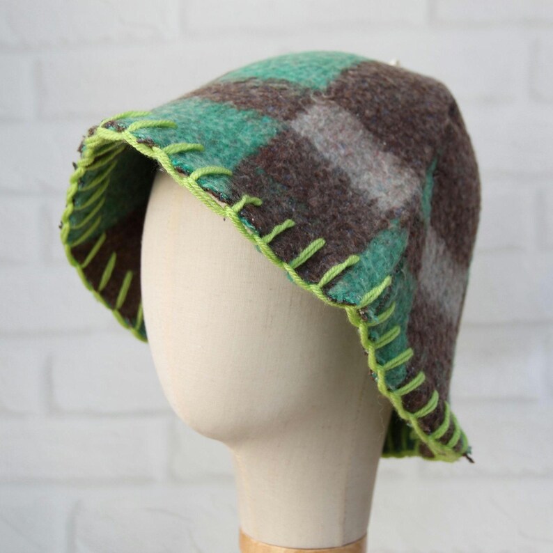 Puede incluir: Un sombrero hecho a mano con un estampado de cuadros en tonos marrones, verdes y grises. El borde est&aacute; adornado con costuras de color verde brillante. El sombrero se exhibe en una cabeza de maniqu&iacute;.
