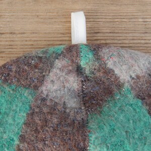 Puede incluir: Primer plano de un sombrero de fieltro con una cinta blanca adjunta. El sombrero presenta un dise&ntilde;o de patchwork en tonos marrones, verde azulado y grises. El sombrero est&aacute; hecho de un material texturizado, similar a la lana.