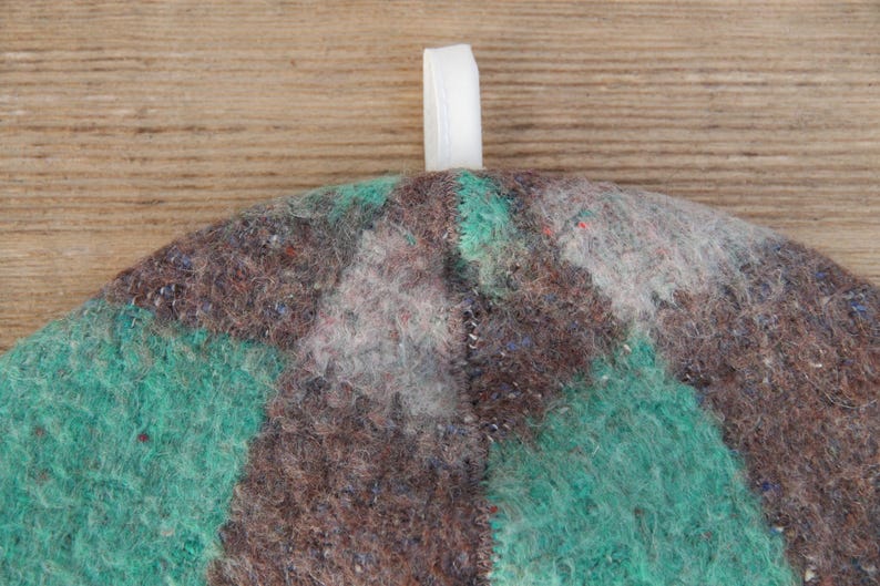 Puede incluir: Primer plano de un sombrero de fieltro con una cinta blanca adjunta. El sombrero presenta un dise&ntilde;o de patchwork en tonos marrones, verde azulado y grises. El sombrero est&aacute; hecho de un material texturizado, similar a la lana.