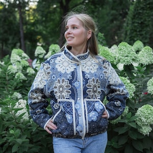 Könnte beinhalten: Eine langärmelige Jacke mit blau-cremefarbenem Blumenmuster, Stehkragen und silbernen Druckknöpfen. Das Kleidungsstück verfügt über zwei Vordertaschen und eine strukturierte Passform, die ein detailliertes Webmuster im Stoffdesign zeigt.