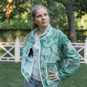 May include: A green and white floral patterned jacket with a round neckline, long sleeves, and a front snap button closure. The jacket features two front pockets and white trim along the edges, worn over a plain white t-shirt and blue jeans.