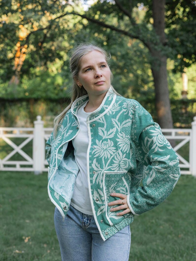 May include: A green and white floral patterned jacket with a round neckline, long sleeves, and a front snap button closure. The jacket features two front pockets and white trim along the edges, worn over a plain white t-shirt and blue jeans.