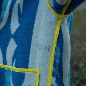 May include: A blue and white patterned blanket coat with yellow trim. The coat features a geometric design in shades of blue and white, with a bright yellow trim outlining the edges and pockets. The fabric appears soft and textured.