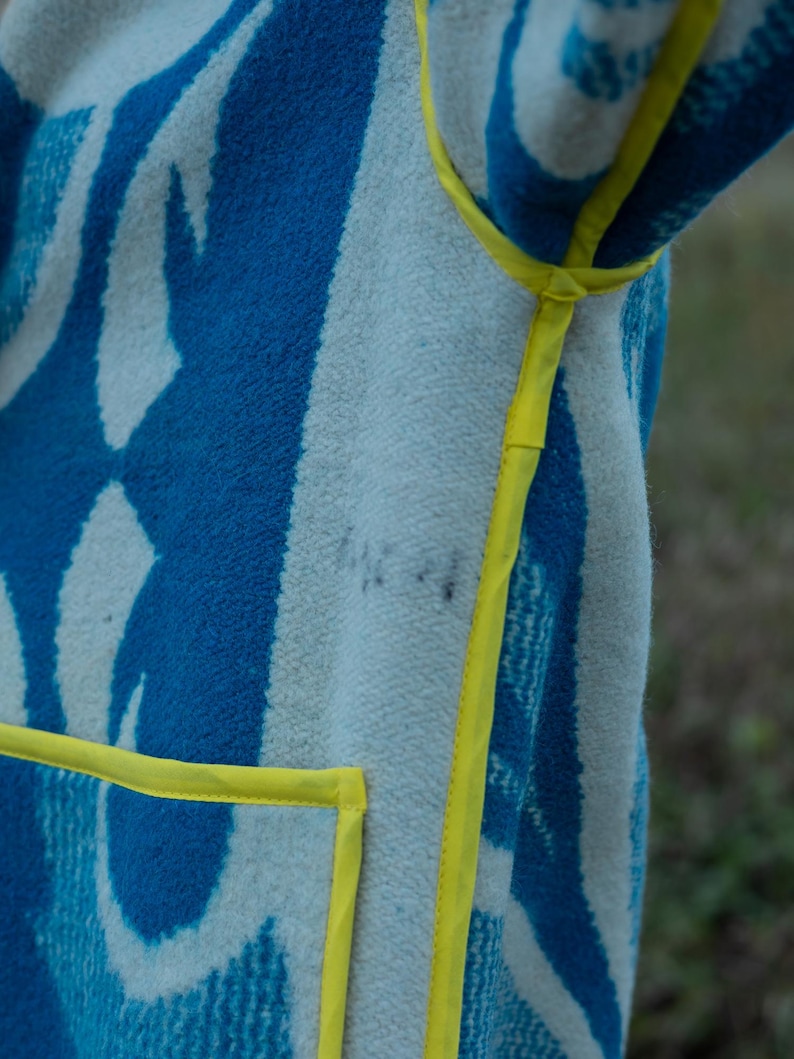 May include: A blue and white patterned blanket coat with yellow trim. The coat features a geometric design in shades of blue and white, with a bright yellow trim outlining the edges and pockets. The fabric appears soft and textured.