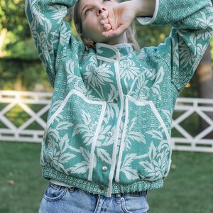 May include: A teal and white floral patterned jacket with a high collar, long sleeves, and white trim along the front closure and pockets. The jacket features silver-toned snap buttons down the center and a textured fabric construction.
