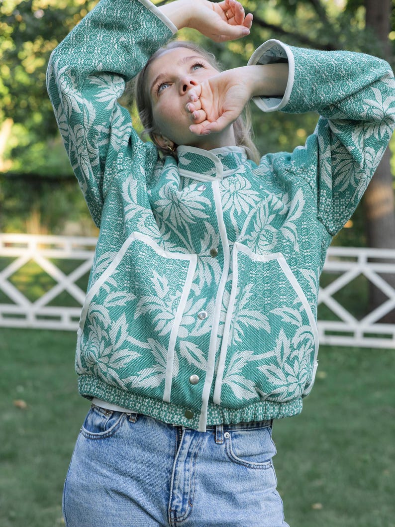 May include: A teal and white floral patterned jacket with a high collar, long sleeves, and white trim along the front closure and pockets. The jacket features silver-toned snap buttons down the center and a textured fabric construction.