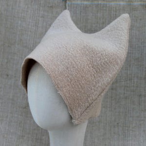 May include: A beige, textured, fleece-like fabric hat with two pointed, cat-ear-shaped peaks at the top. The hat features a structured, rounded front opening and visible stitched seams along the edges, crafted from a soft, fuzzy material.