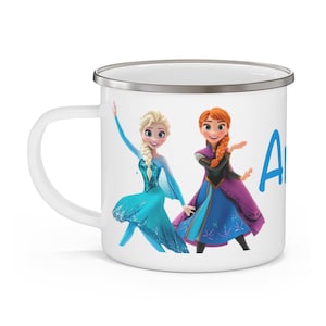 May include: White enamel mug featuring the characters Elsa and Anna from the movie Frozen. Elsa is in a blue dress, and Anna is in a purple and blue dress. The name "Anna" is written in blue on the right side of the mug.