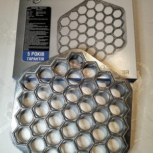 May include: A silver metal pelmeni maker with 36 individual molds for making dumplings. The maker is shaped like a hexagon and has a raised rim around each mold. The maker is sitting on a white surface. The box for the pelmeni maker is in the background and has the text "Пельменница" on it.