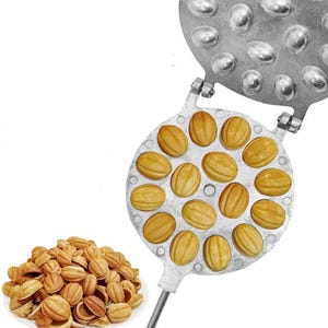 May include: A metal walnut-shaped cookie maker with a hinged design, open to reveal baked cookies. A pile of finished, golden-brown walnut cookies sits on a white plate, ready to be filled. The baking tool is silver.