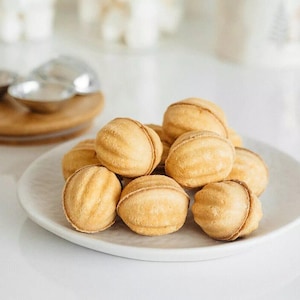 May include: A white plate with a pile of golden brown walnut-shaped cookies. The cookies are arranged in a pyramid shape and have a slightly textured surface.