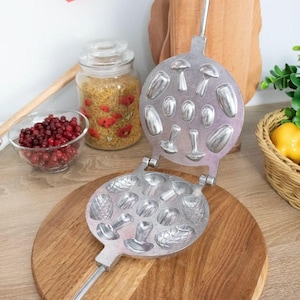 May include: A silver waffle iron with a wooden handle. The iron has two sections, one with mushroom and acorn shapes and the other with mushroom shapes. The iron is open and sitting on a wooden surface.