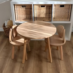 Op de afbeelding: Een lichtgekleurde houten kindertafel met een ronde bovenkant en twee bijpassende stoelen. De stoelen hebben gebogen rugleuningen en poten. De tafel en stoelen staan op een hardhouten vloer. Een witte plank met geweven manden staat op de achtergrond.
