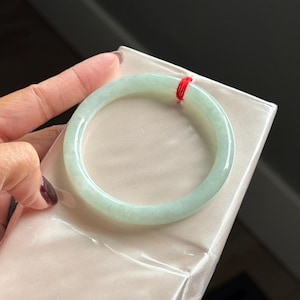 May include: A smooth, pale green translucent bangle bracelet with a polished finish. The circular jewelry piece features a thin, vibrant red string tied around one section of the band, resting against a smooth, light-colored satin-like surface.