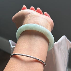 May include: A smooth, pale green jade bangle bracelet worn on the wrist alongside a thin, polished silver-tone metal bangle featuring a repeating textured pattern. The jade bangle has a translucent appearance with subtle variations in its light green color.