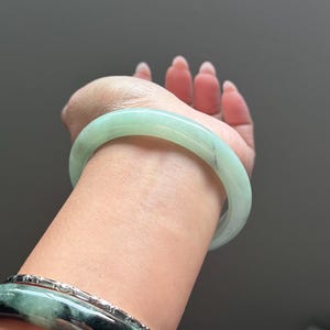 May include: A smooth, translucent pale green jade bangle bracelet worn on the wrist, accompanied by a second bangle featuring a mottled pattern of dark green and black swirls with a metallic silver-toned textured band layered alongside it.