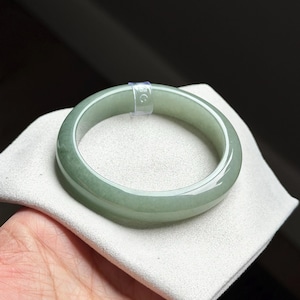 May include: A smooth, polished, light green jade bangle bracelet with a translucent appearance. The circular bangle features a small, clear plastic tag attached to the top edge, displaying the number 59 in white text.