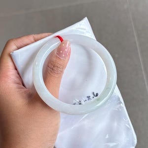 May include: A smooth, translucent white jade bangle bracelet with a polished finish. The circular band features a subtle, faint green tint and a small red string loop attached to the top. The number 58.6 is written in black ink on the inner surface.