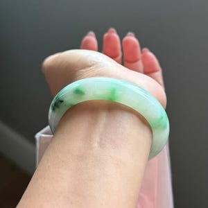 May include: A smooth, polished jade bangle bracelet featuring a translucent white base with vibrant green patches and subtle dark green inclusions. The circular accessory has a rounded profile and a glossy finish, showcasing natural color variations.