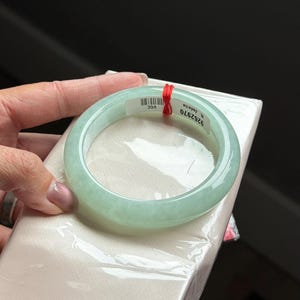 May include: A smooth, translucent pale green jadeite bangle bracelet with a rounded profile. A white adhesive label is attached to the inner surface, featuring the text 9262970, N. Jadeite, and VGC, secured by a thin red string.