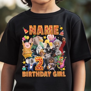 May include: Black t-shirt featuring a colorful graphic of animated characters, the number "2", and the text "NAME" and "BIRTHDAY GIRL" in a marquee-style font. The design includes stars, balloons, and a party hat.