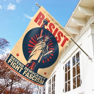 May include: A beige flag with the word "RESIST" in red, and the Statue of Liberty in the center. The flag also has the text "DEFEND DEMOCRACY FIGHT FASCISM" in a blue box with red stars. The flag is hanging outside a white building.