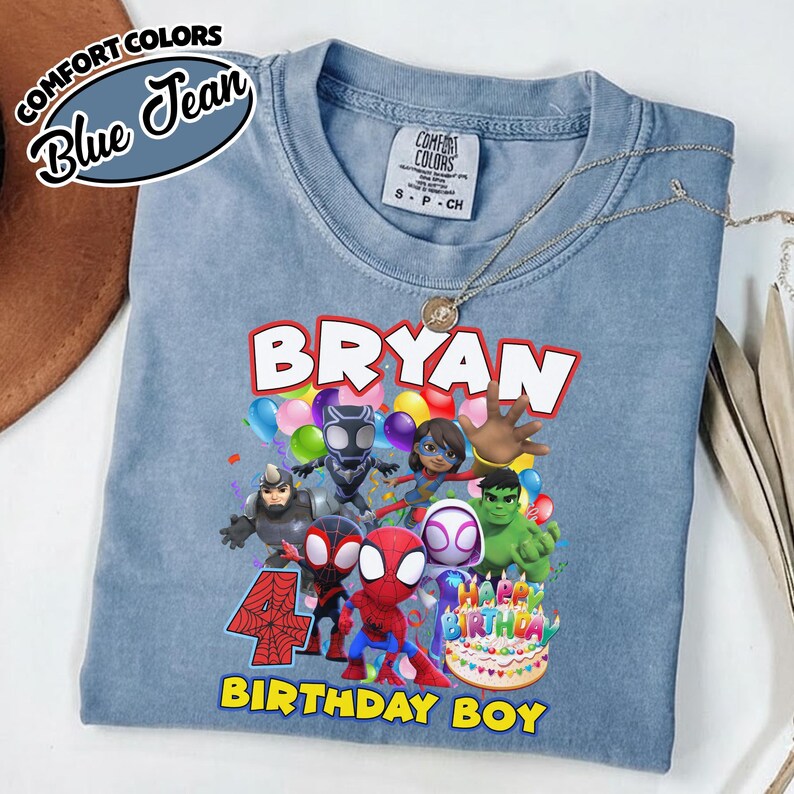 May include: A blue jean colored t-shirt featuring a graphic print of various superhero characters, colorful balloons, a birthday cake, and the text "BRYAN" at the top and "4 BIRTHDAY BOY" at the bottom. The shirt has a Comfort Colors brand label.