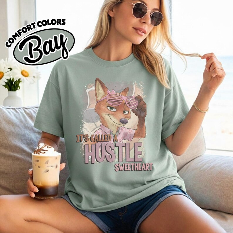 Puede incluir: Una camiseta de manga corta color verde salvia con un gr&aacute;fico de un zorro de dibujos animados con gafas de sol rosas de persiana y una camisa estampada. El texto dice "IT'S CALLED A HUSTLE SWEETHEART". La camiseta est&aacute; marcada como "COMFORT COLORS" y "BAY".