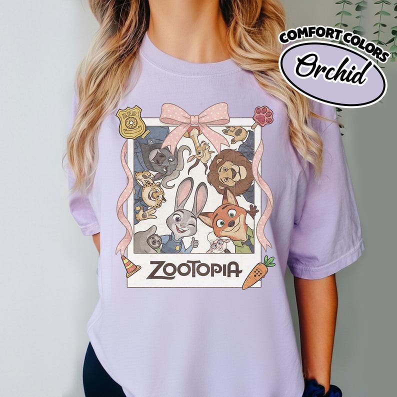 May include: A light purple short-sleeve t-shirt featuring a graphic print of Zootopia characters inside a white polaroid-style frame. The design includes a pink bow, a police badge, a paw print, a traffic cone, and a carrot with the text Zootopia.