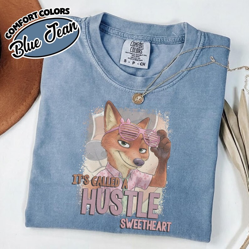 Puede incluir: Una camiseta azul de manga corta con un gr&aacute;fico de un zorro que lleva gafas de sol de persiana rosas. El zorro viste una camisa rosa claro estampada. Debajo del gr&aacute;fico, el texto dice: IT'S CALLED A HUSTLE SWEETHEART. La camiseta tiene una etiqueta Comfort Colors.