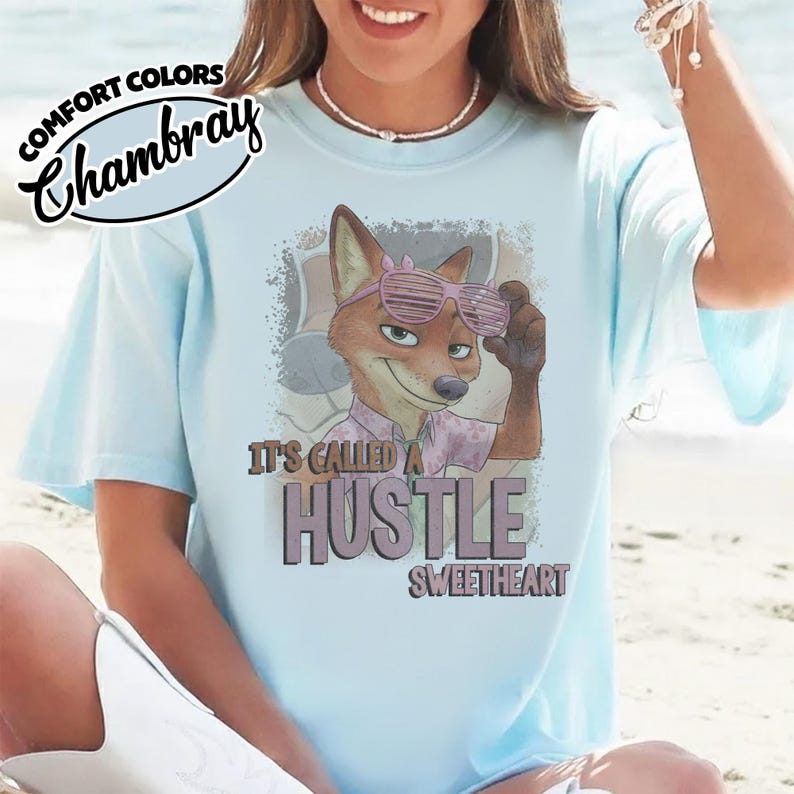 Puede incluir: Una camiseta azul claro con un gr&aacute;fico de un zorro de dibujos animados con gafas de sol de persiana rosas. El texto dice "IT'S CALLED A HUSTLE SWEETHEART" en negrita. Un logotipo circular en la esquina superior izquierda muestra "COMFORT COLORS Chambray".