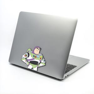 K&ouml;nnte beinhalten: Ein farbenfroher Aufkleber von Buzz Lightyear aus Toy Story ist auf der R&uuml;ckseite eines silbernen Laptops angebracht. Der Aufkleber zeigt Buzz in seiner Weltraumranger-Uniform mit gr&uuml;nen, wei&szlig;en, lila und roten Akzenten. Der Laptop ist ge&ouml;ffnet und zeigt die Tastatur.