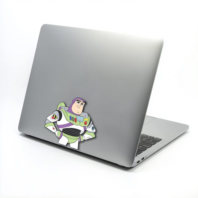 K&ouml;nnte beinhalten: Ein farbenfroher Aufkleber von Buzz Lightyear aus Toy Story ist auf der R&uuml;ckseite eines silbernen Laptops angebracht. Der Aufkleber zeigt Buzz in seiner Weltraumranger-Uniform mit gr&uuml;nen, wei&szlig;en, lila und roten Akzenten. Der Laptop ist ge&ouml;ffnet und zeigt die Tastatur.